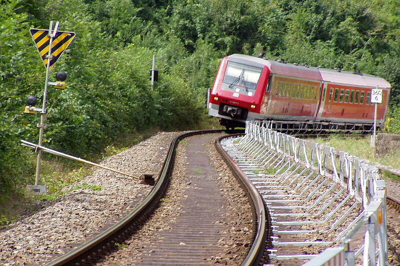 Heranfahrender roter Zug im Gleis mit automatischem Warnsystem und festem Absperrungszaun.