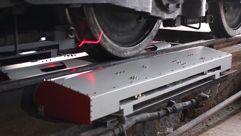 Ein Foto, das eine metallische, im Gleis eingebettete Box zeigt. Darüber sind das Räderwerk eines Zugs zu sehen und der rosafarbene Laserstrahl, der aus der Box kommt.